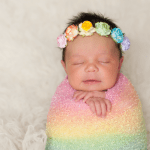 Baby laying on blanket wrapped in rainbow swaddle and wearing a flower headband