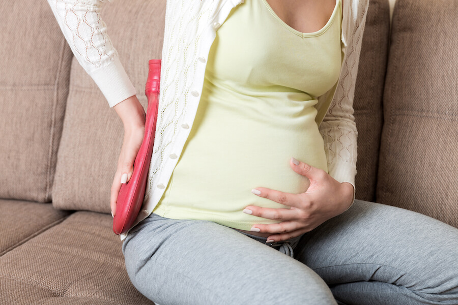 A photo of a pregnant woman on the couch, holding a hot water pack on her back with one hand and her other hand on her stomach.