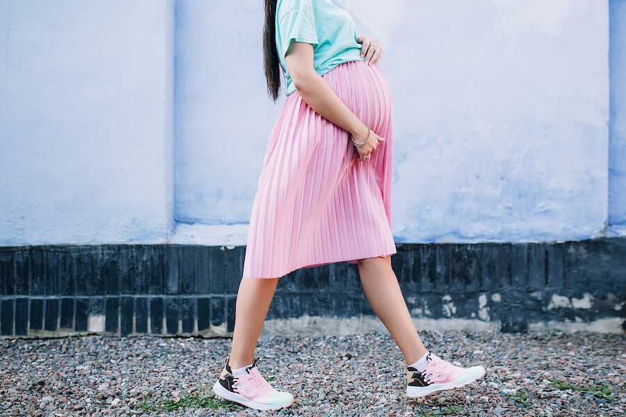 A photo of a pregnant woman walking down the street in a long light pink skirt and light blue shirt while holding her stomach.