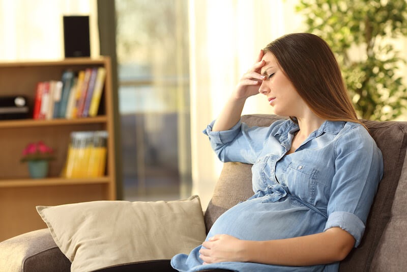 A pregnant woman is sitting on the couch holding her belly with one hand and her forehead with the other