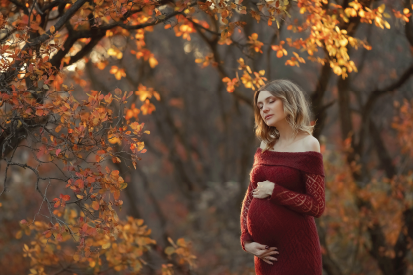Pregnant woman in a maroon dress, holding her bump in a field of trees posed for a fall maternity shoot.