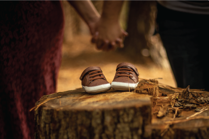 Baby shoes on a tree stump in the foreground with a blurred out man and woman holding hands in the background.