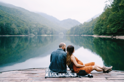 Man and pregnant woman sitting on a deck together with a lake and mountains behind them.